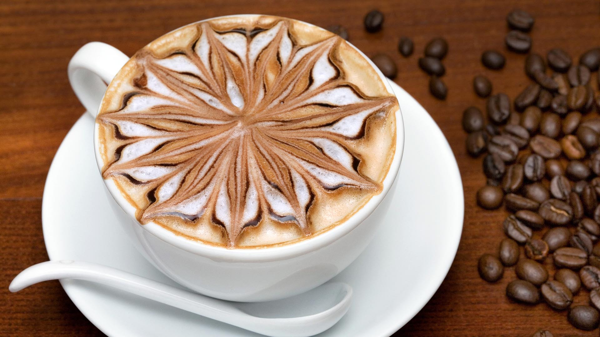 Filiżanka cappuccino z artystycznym wzorem na mlecznej piance, obok rozsypane ziarna kawy.